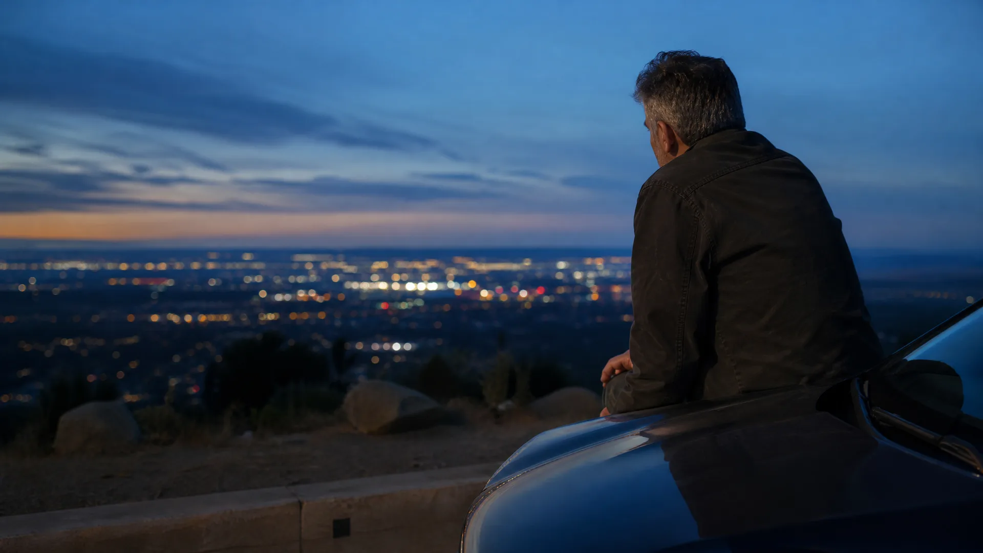 A man in a dark jacket leans against a car on a hillside overlook at dusk, facing away toward distant city lights below a blue sky.